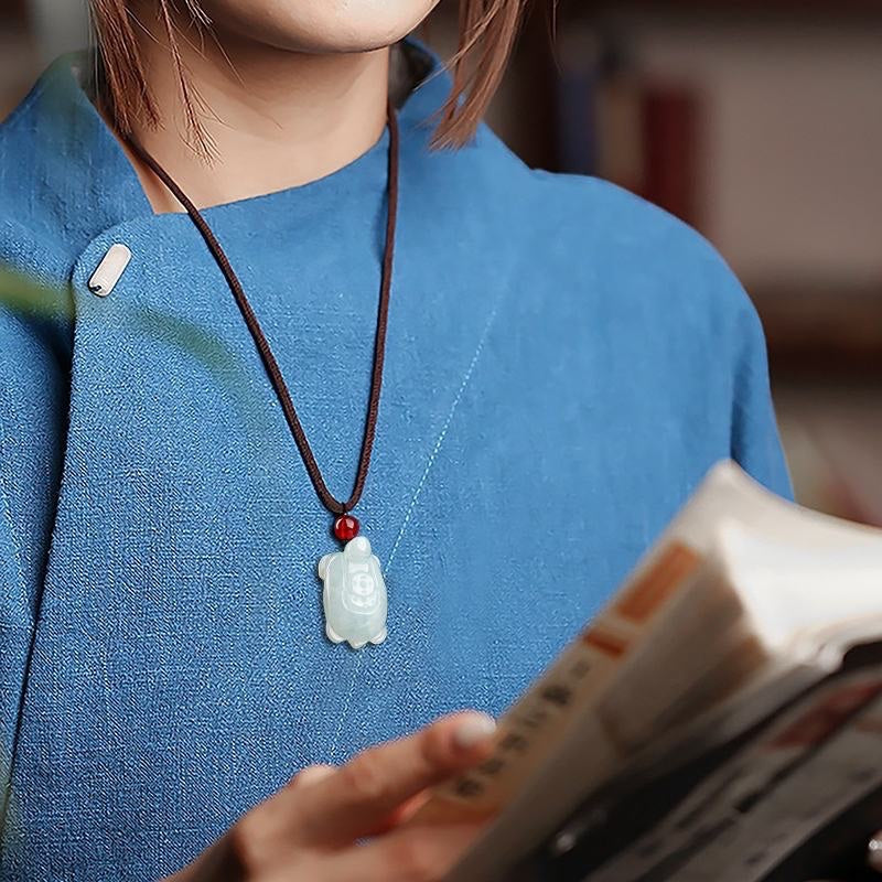 BoxUnboxed hand-carved natural jadeite turtle pendant on a brown cord worn by a person in a blue linen top.